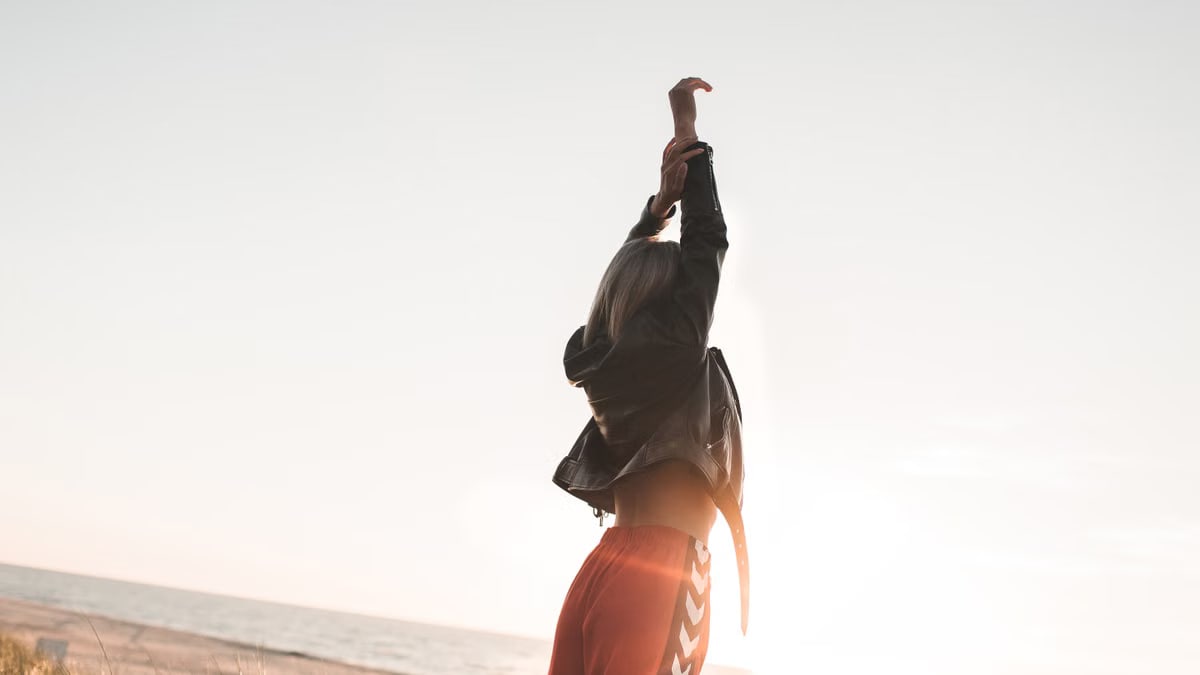 A fashionable woman stands with arms raised on a beach at sunset, exuding style and freedom.