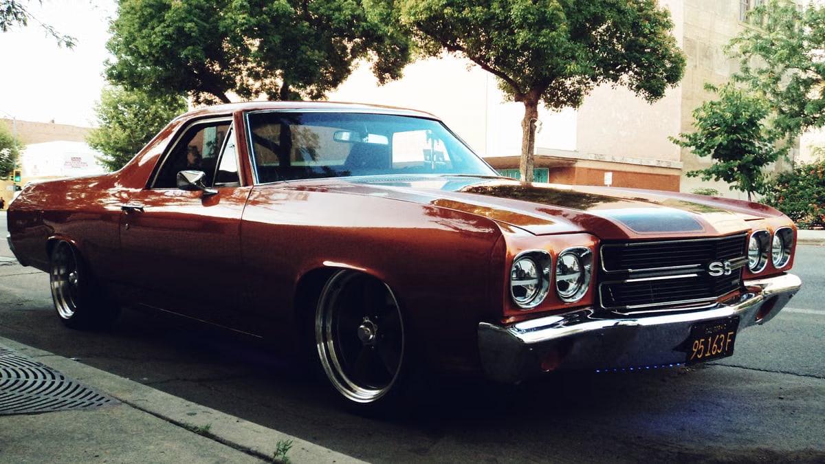 Red Chevrolet El Camino parked on a tree-lined city street, evoking a nostalgic and retro feel.